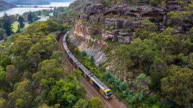 A train on tracks pictured from above with a sandstone wall and water in the background