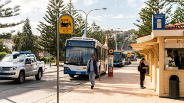 A line of busses waiting at stops, with people walking on the foot path and trees in the background