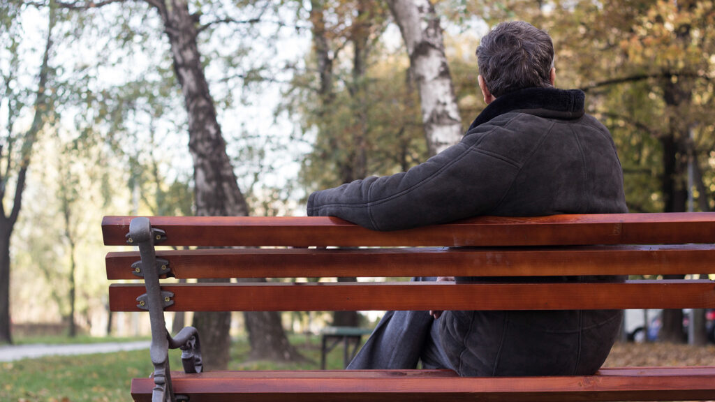 A dark haired man wearing a black jacket sitting on a park bench, pictured from behind