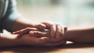 Two hands holding another person's one hand resting on a wooden table