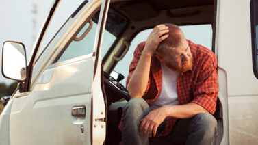 Man wearing red flannel shirt and black jeans sitting in the drivers seat of a white van with one hand resting on his head