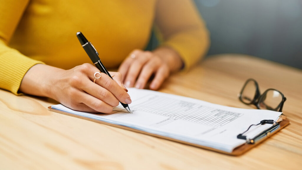 A woman's torso pictured from the shoulders down wearing a yellow jumper, signing some paperwork on a wooden table