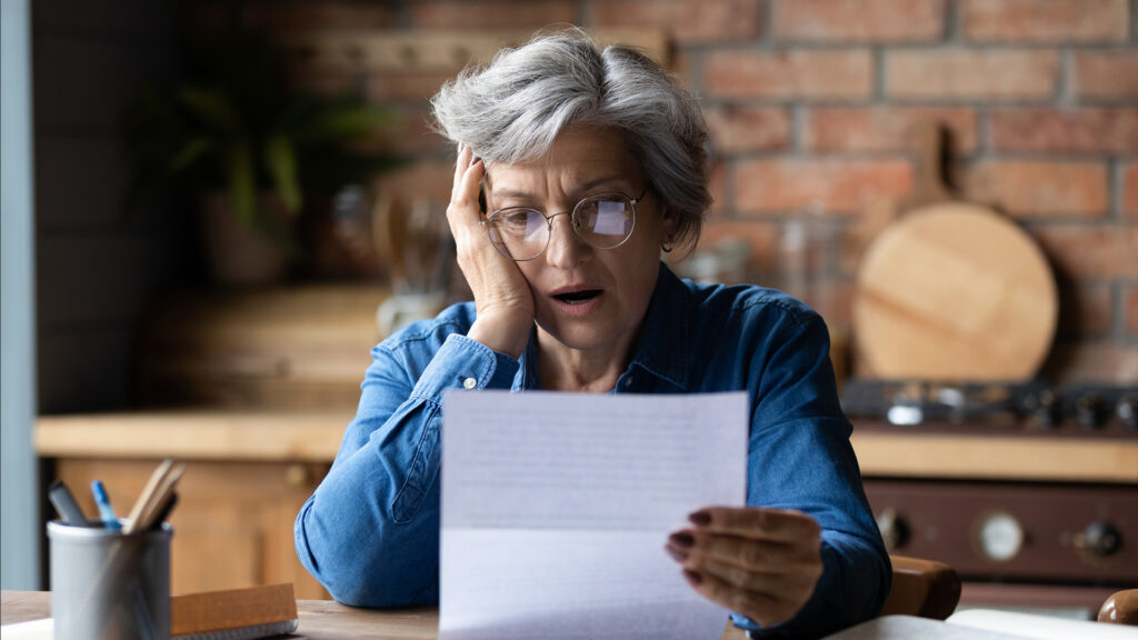 Woman with grey hair wearing glasses looks shocked by the document she's holding