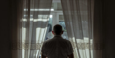 Rear view of adult man standing beside window with sunlight and shadow